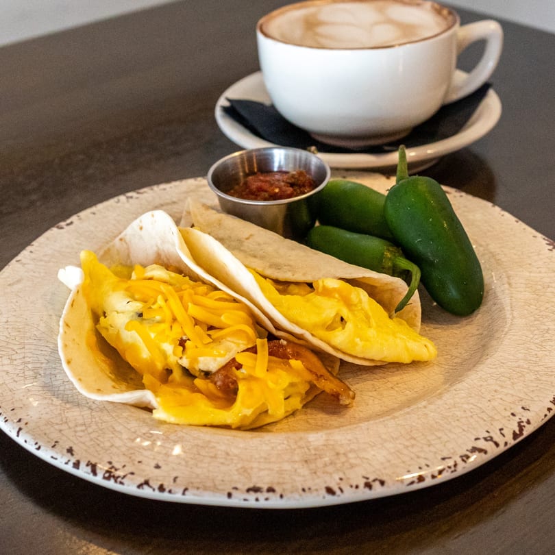 Breakfast tacos with scrambled eggs, cheese, salsa, jalapeños, and a latte on a rustic plate.