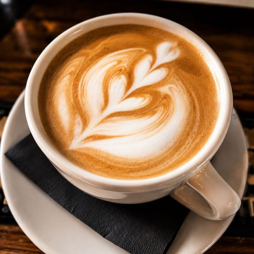 A white cup of latte with intricate foam art, served on a saucer with a black napkin.