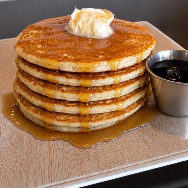 Stack of five golden pancakes with melting butter and syrup, next to a metal cup of syrup.