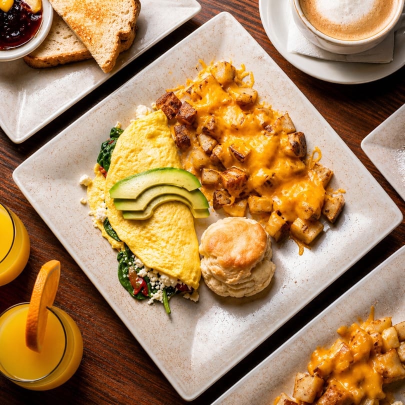 Breakfast plate with omelet, avocado, biscuit, cheesy potatoes, toast, coffee, and orange juice.