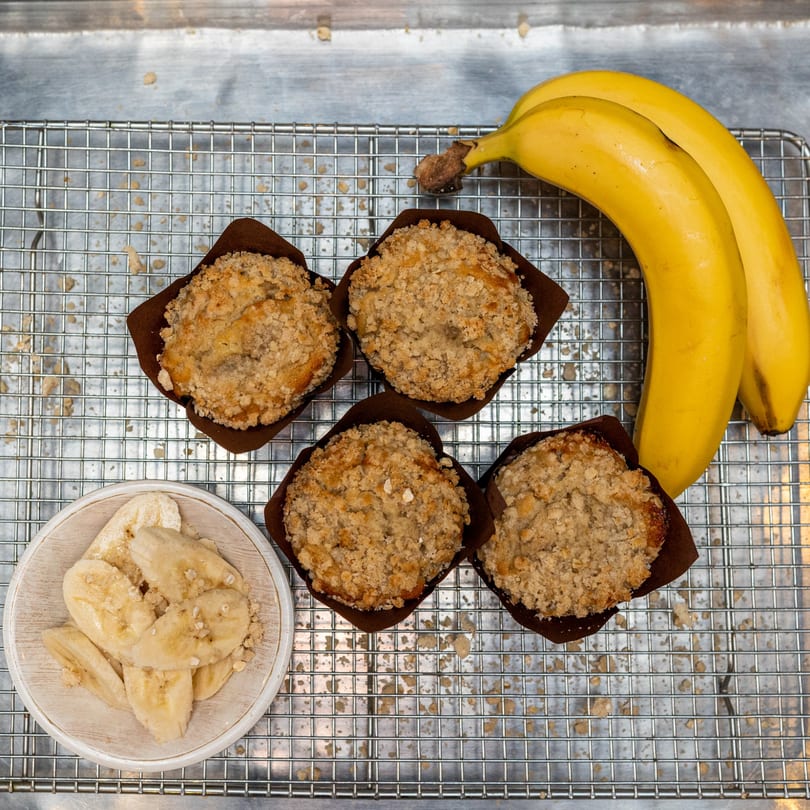 Four banana muffins with crumble topping, two bananas, and sliced bananas on a cooling rack.