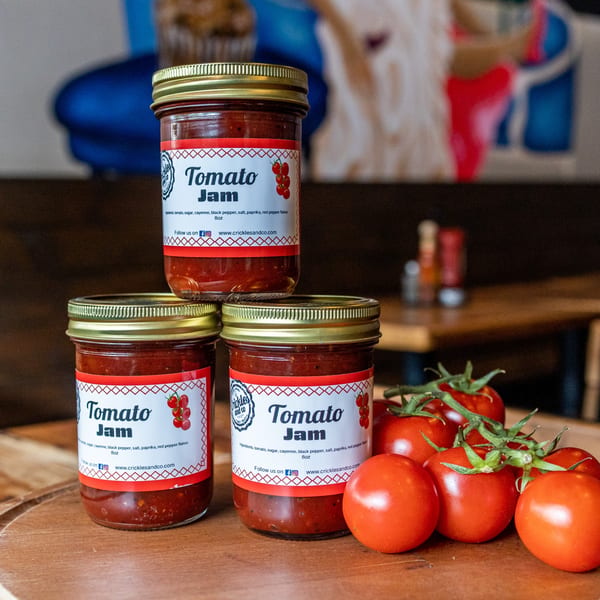 Three jars of Crickles and Co. Tomato Jam and a cluster of fresh red tomatoes on a wooden surface.