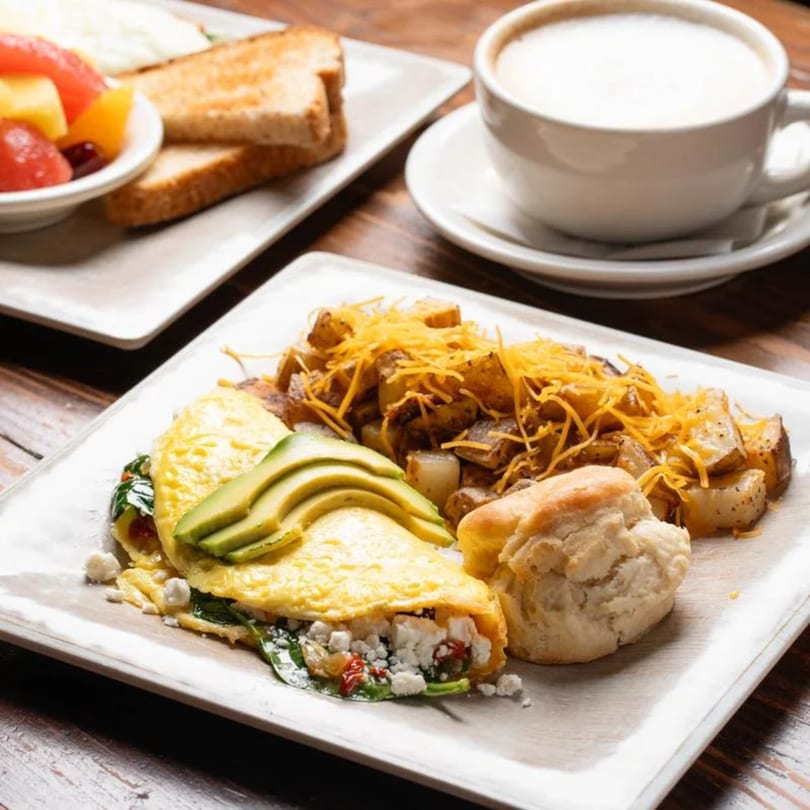 A breakfast plate with an avocado omelet, cheesy potatoes, biscuit, fruit cup, and coffee.
