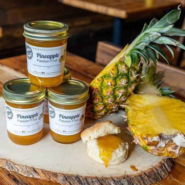 Jars of pineapple passion fruit jam, a halved pineapple, and a biscuit with jam on a wooden board.