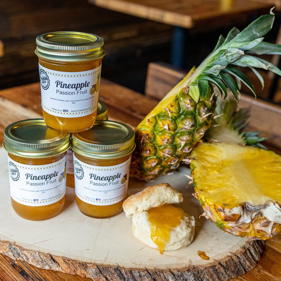 Jars of pineapple passion fruit jam, a halved pineapple, and a biscuit with jam on a wooden board.