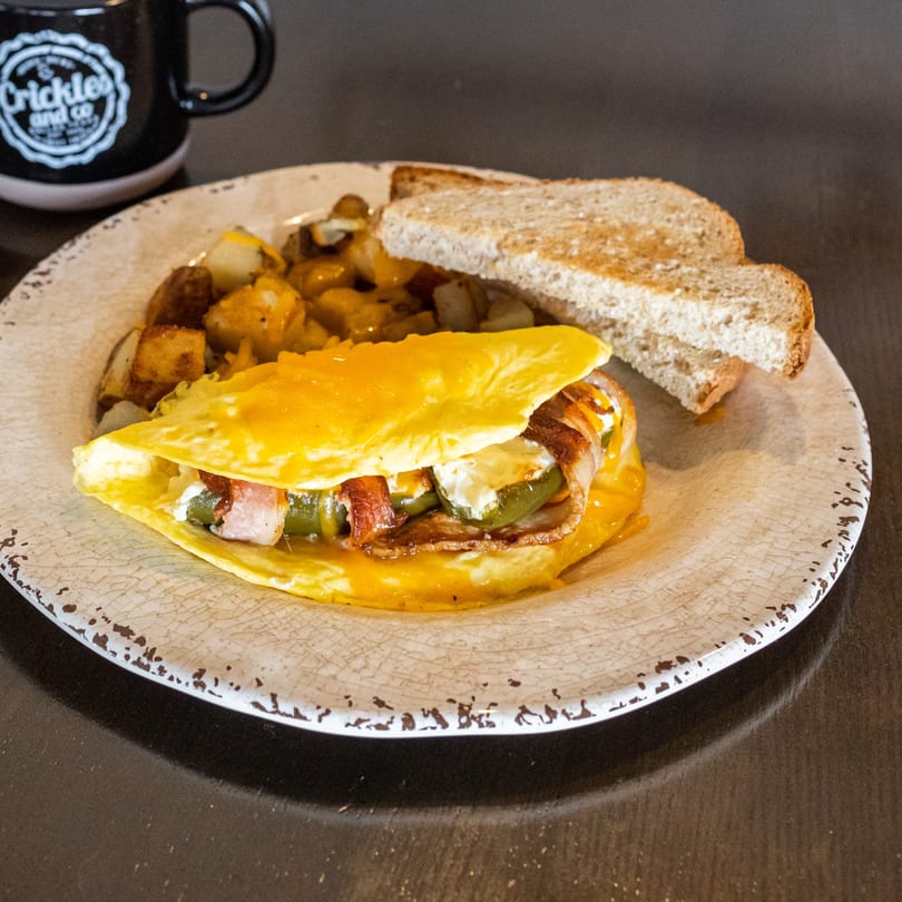 A cheesy omelet with bacon and jalapeños, served with home fries and toast, next to a coffee mug.
