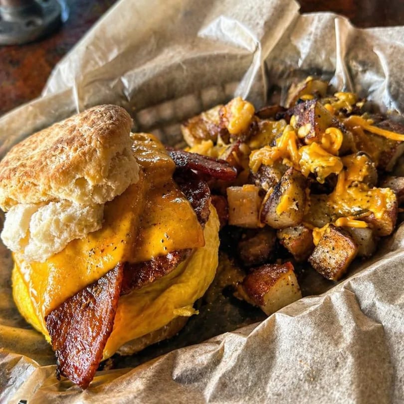 A breakfast biscuit sandwich with bacon, egg, and cheese, served with seasoned potatoes.
