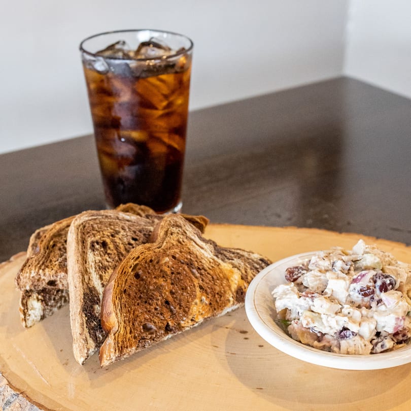 Chicken salad with grapes and pecans, toasted bread, and a glass of iced soda on a wooden board.