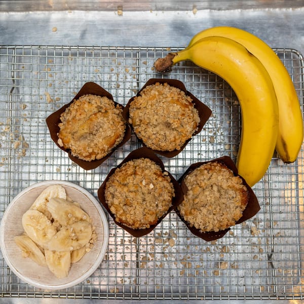 Four banana crumb muffins, two bananas, and a bowl of sliced bananas on a cooling rack.